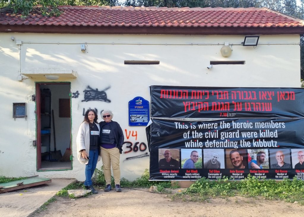 Liora und Hadas vor einem Plakat mit Menschen, die bei der Verteidigung des Kibbutz ums Leben kamen
